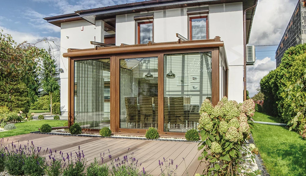 Ein modernes Haus mit einem Wintergarten mit großen Glasfenstern und braunem Rahmen, umgeben von einem Garten mit Blumen, Sträuchern und einer Holzterrasse.