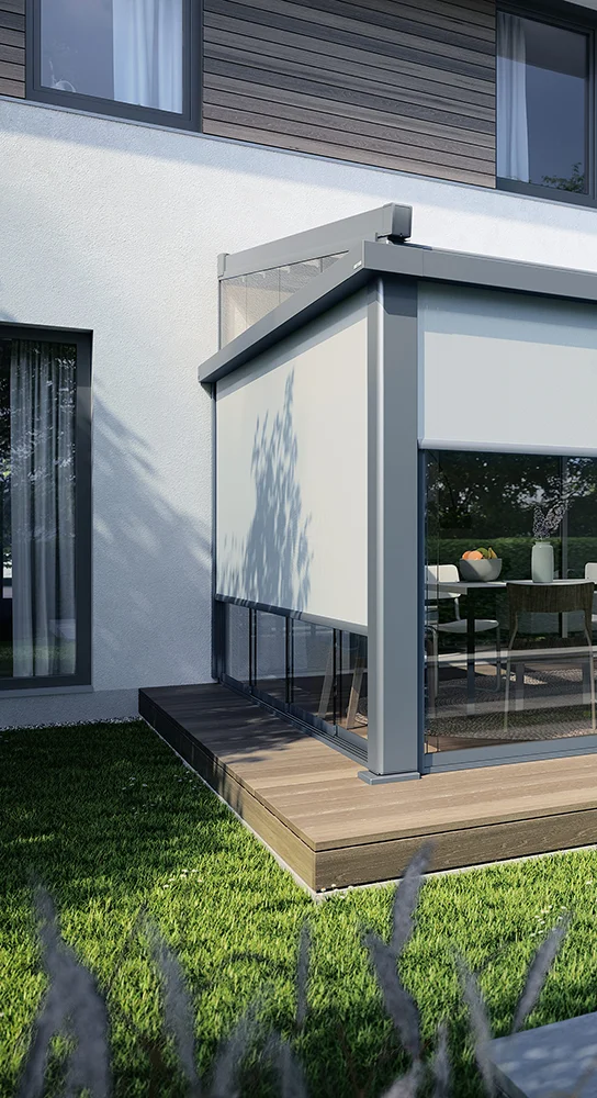 Eine moderne Außenterrasse mit Glaswänden und einem heruntergelassenen weißen Rollladen befindet sich auf einer Holzterrasse, die an ein modernes Haus angebaut ist, umgeben von grünem Gras und teilweise im Schatten von Bäumen.