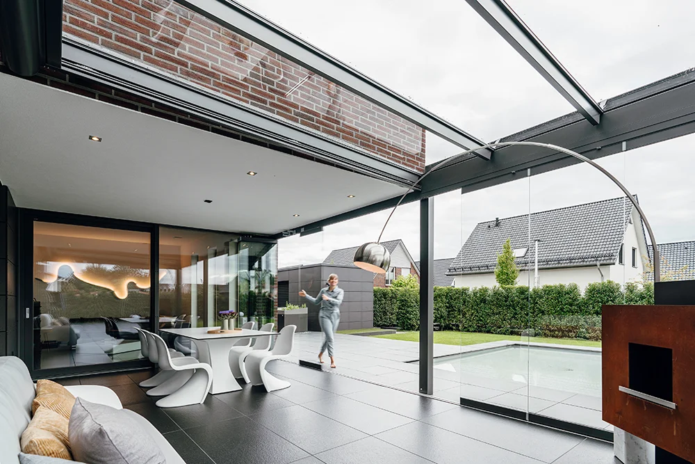 Ein moderner Innenhof mit Glaswänden und -decken, stilvollen weißen Möbeln und einem Pool. Eine Person geht auf der Terrasse spazieren, und im Hintergrund sind moderne Häuser und Grünflächen zu sehen.