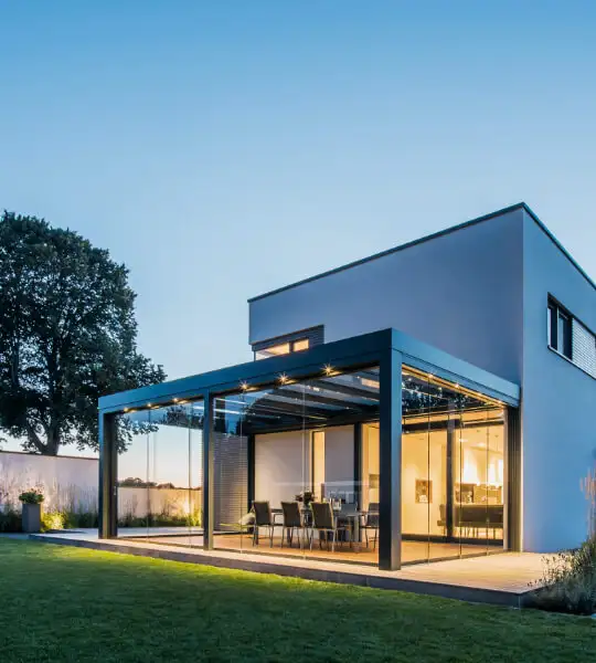 Modernes zweistöckiges Haus mit großen Glasfenstern und einer überdachten Terrasse, einem Essbereich im Freien und einem gepflegten Rasen bei Sonnenuntergang. Ein großer Baum steht an der Seite, und der Himmel ist klar.